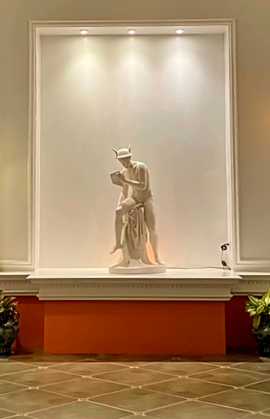 Statue of a seated figure on a pedestal against a white wall with an orange base.