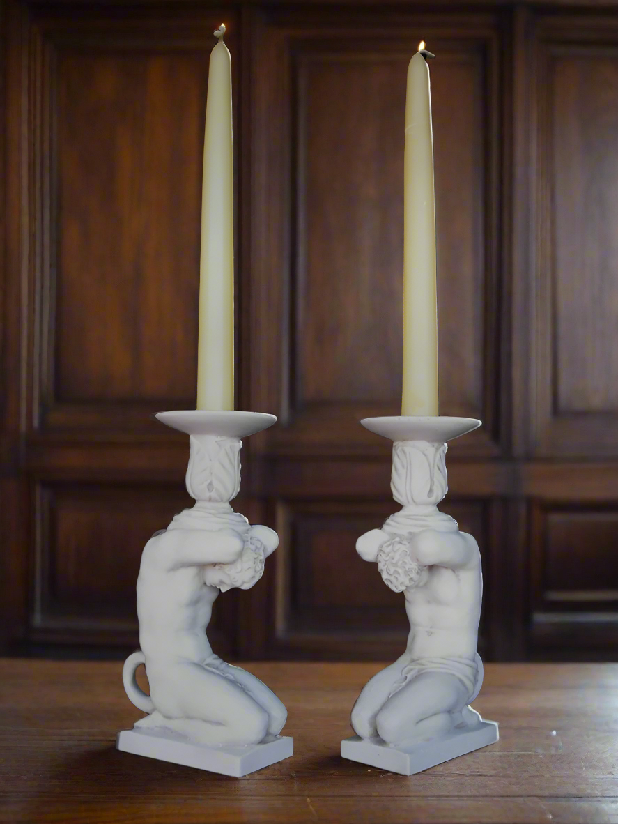 Two white candle holders shaped like classical statues with candles on a wooden surface.