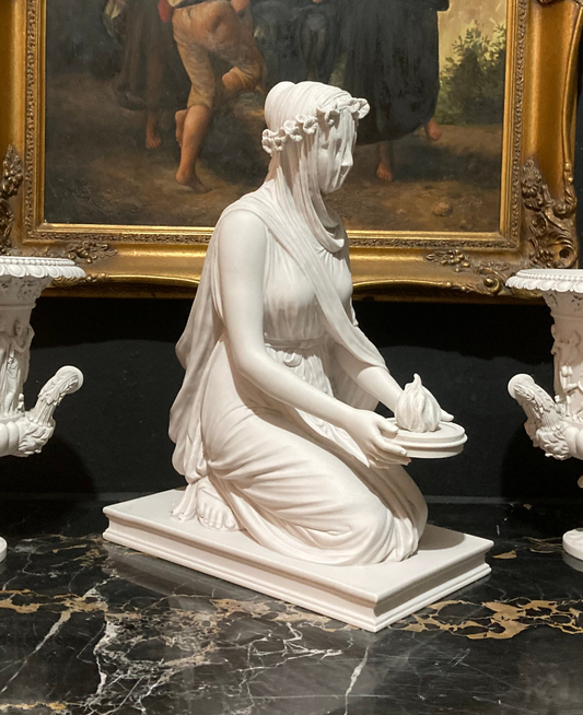 White marble statue of a woman holding a candle on a dark marble surface with a painting in the background.