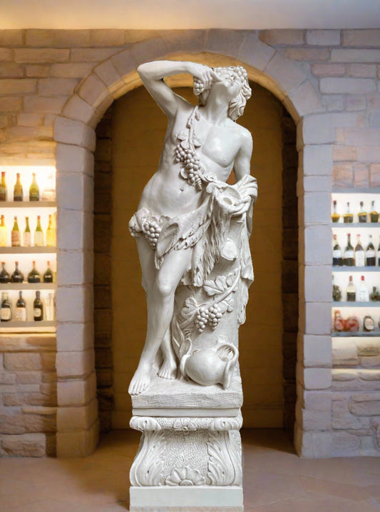 A marble statue of Bacchus, depicting the Greek god Dionysus, with a goblet of wine and an empty wine ewer at his feet, standing on an ornate plinth with grapes on the vine. The statue is displayed in a room with stone walls and a wine bottle collection.