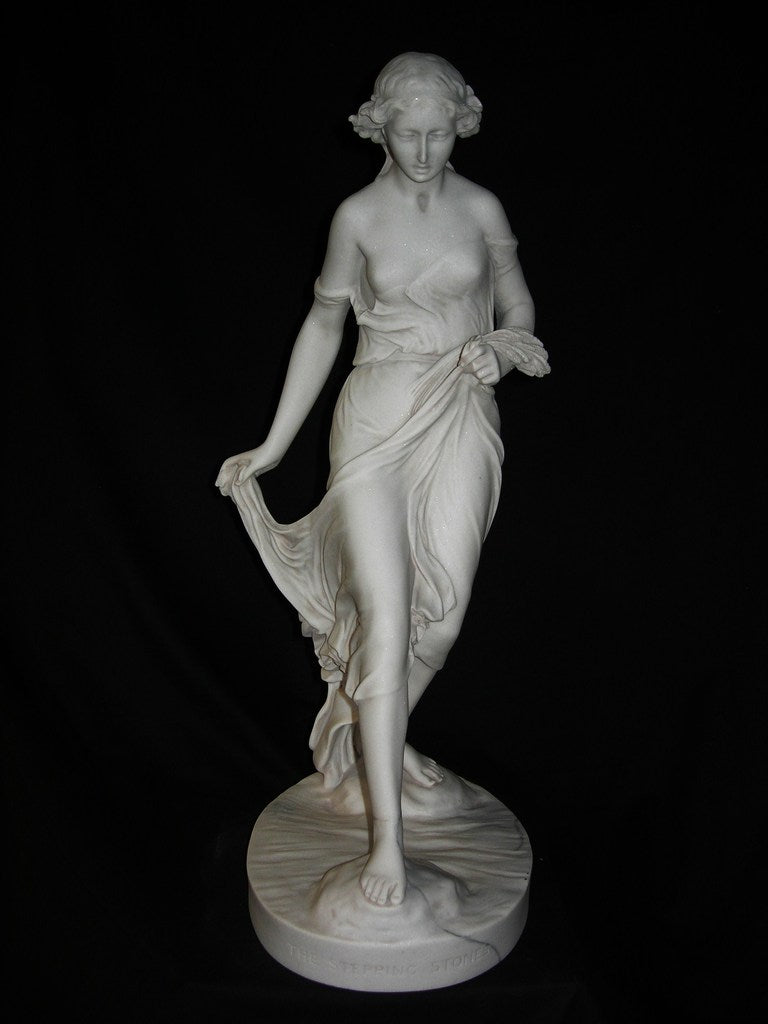 A white marble sculpture of a female figure standing on one foot, with a flowing garment, on a circular base. The sculpture is illuminated from above, highlighting its details against a dark background.
