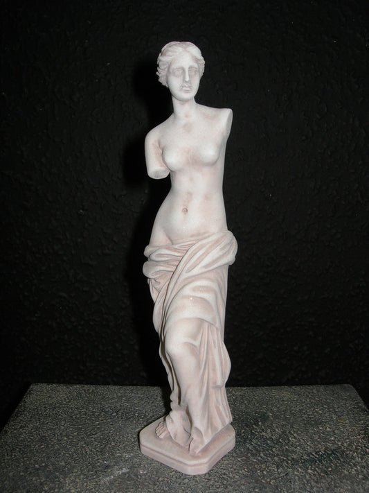 A miniature statue of Venus de Milo, depicting a nude female figure with flowing drapery, primarily in a pink or light color, set against a black background.