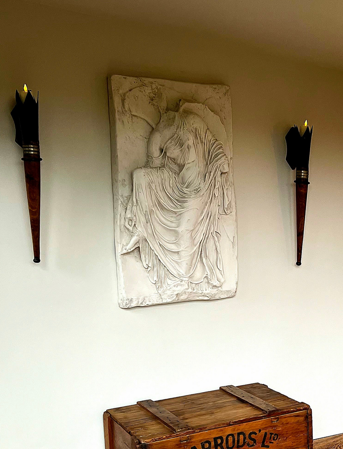 Decorative wall piece with a seated figure between two torches on a plain wall.