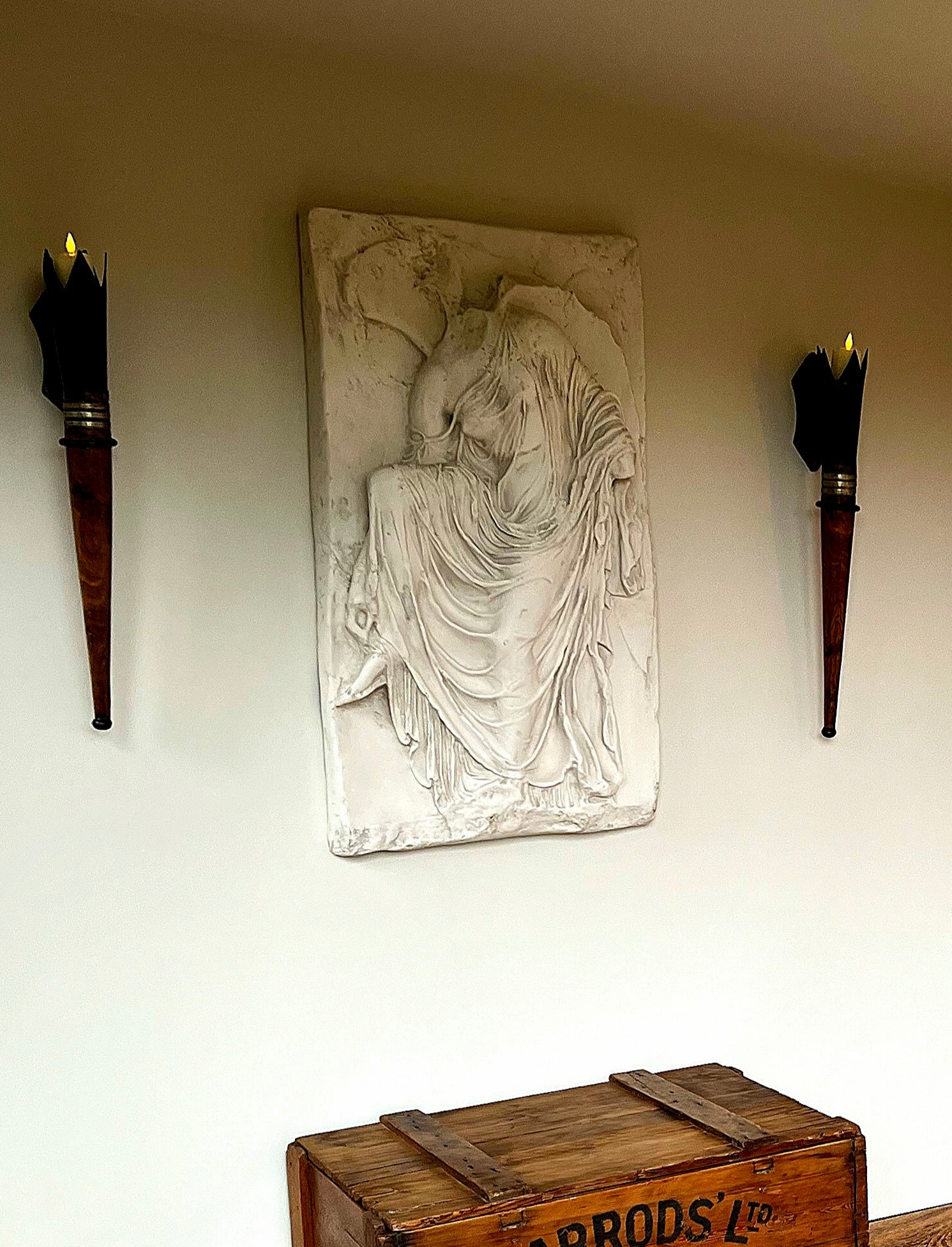 Decorative wall piece with a seated figure between two torches on a plain wall.