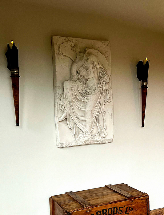 Decorative wall piece with a seated figure between two torches on a plain wall.