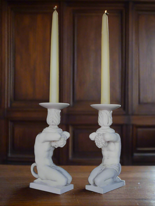 Two white candle holders shaped like classical statues with candles on a wooden surface.